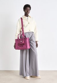 Różowa torba z płótna z uchwytami i paskiem na ramię, z napisem "Coach New York". Modelka ma na sobie off-white pikowaną kurtkę i szare spodnie o szerokich nogawkach.