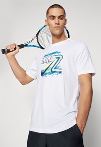 Valge T-särk, millel on sinise ja rohelise graafikaga logo. Mudel hoiab tennisereketit, lühikeste varrukatega ja lõdva lõikega.