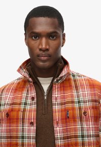 Chaqueta de cuadros en naranja, rojo, blanco y verde, con un suéter marrón con cremallera por debajo. Cuenta con un cuello alzado y cierres de botones.