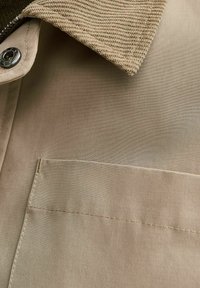 Beżowa tkanina z mieszanki bawełny z teksturowanym kołnierzem sztruksowym. Posiada kieszeń z klapą oraz widoczne detale szwów. Gładka i matowa faktura.