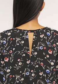 Blusa nera floreale con un taglio a V sulla schiena. Presenta un motivo di fiori rossi, blu e bianchi su un tessuto testurizzato.