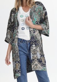 Kimono-lik jacka i marinblått, grönt och beige paisleymönster, lättviktig tyg, öppen fram, lös passform, tillsammans med en vit grafisk t-shirt och jeans.