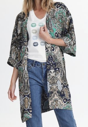 Kimono-stijl jas in navy, groen en beige paisleydessin, lichtgewicht stof, open voorkant, losse pasvorm, gecombineerd met een wit graphic t-shirt en jeans.