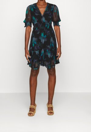 Žena v krátkých černých šatech se zeleno-modrým květinovým vzorem, s nafouknutými průsvitnými rukávy, výstřihem do V a béžovými otevřenými sandály na podpatku.