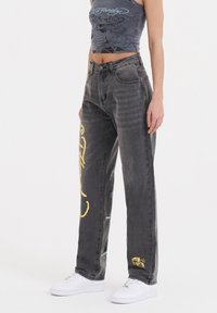 Sorte denim jeans med en falmet finish, prydet med gule grafiske designs på venstre ben og en rullet kant. Sammen med en grå mønstret crop top og hvide sneakers.