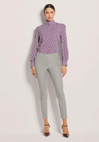 Frau in einer lila gemusterten Bluse mit hohem Kragen, hellgrauen schmalen Hosen und dunkelbraunen spitzen Schuhen, die gegen eine schlichte Wand steht.