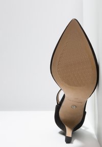 Chaussure à talon haut en daim noir avec un bout pointu, un talon fin et une semelle beige lisse. Dotée d'une bride pour le maintien de la cheville.