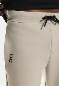 Beige Jogginghosen aus weichem Stoff, mit einem Tunnelzugbund und seitlichen Taschen. Schwarzer Logodruck auf dem linken Bein.