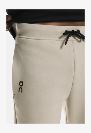 Pantalones jogger beige hechos de tejido suave, con cinturilla ajustable y bolsillos laterales. Estampado del logo en negro en la pierna izquierda.