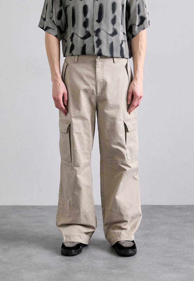 Pantalons cargo beiges avec poches latérales, design à jambes larges, fabriqués en tissu durable. Associés à une chemise à boutons noir et blanc à motif.