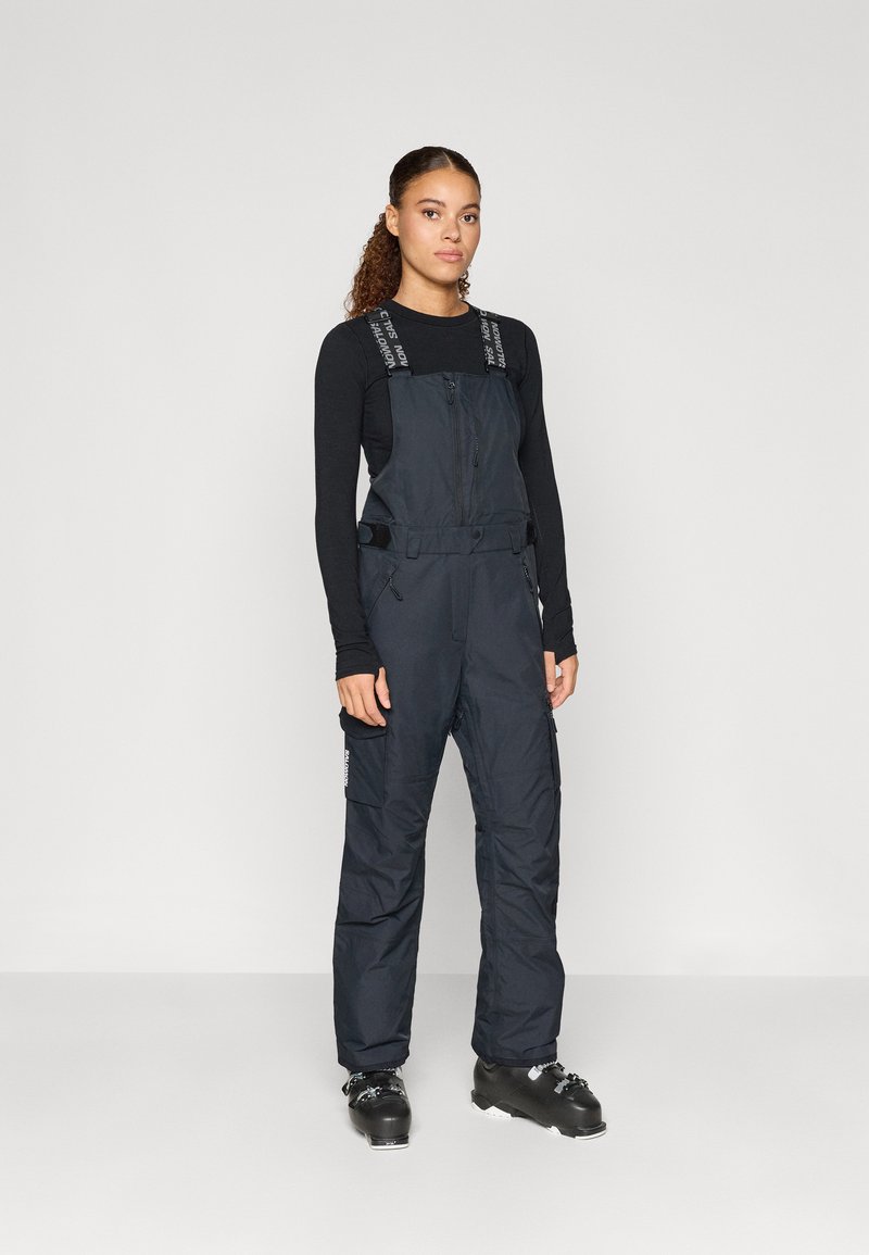 Fekete szigetelt síoverall állítható pántokkal, elöl cipzárral és oldalsó zsebekkel, fekete sícipőkkel párosítva. Simább textúra.