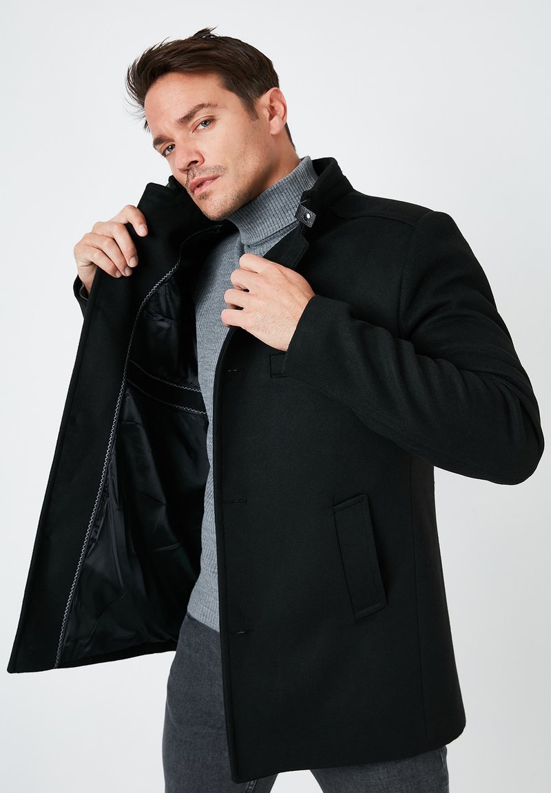 Schwarzer Wollmantel mit Kragen und Knopfverschluss, mit einem glatten Innenfutter. Kombiniert mit einem hellgrauen Turtle-Neck.