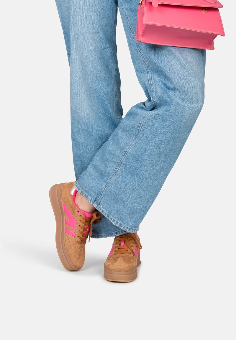 Baskets en suède marron avec des accents et des lacets roses, dotées d'une semelle épaisse beige. Portées avec un jean bleu à pattes larges.