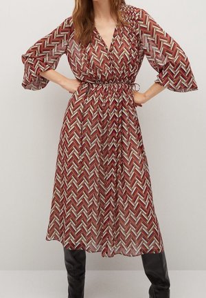 Kobieta ubrana w sukienkę za kolano z długim rękawem, o czerwono-beżowo-brązowym wzorze tkanym, w połączeniu z czarnymi, wysokimi do kolan kozakami.