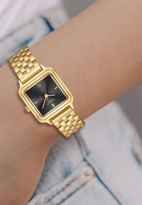 Zelts taisnstūrveida pulkstenis ar melnu seju, ar teksturētu metāla siksniņu un diviem maziem stundu rādītājiem. Novietots uz plaukstas, kur redzama ādas krāsa.