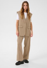 Beige tailored vest with wide armholes, paired with loose-fitting beige trousers. Worn over a white shirt, complemented by black platform shoes.