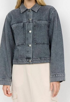 Femme portant une veste en jean noire délavée avec fermeture à boutons et grandes poches avant, associée à un pantalon cargo beige clair.