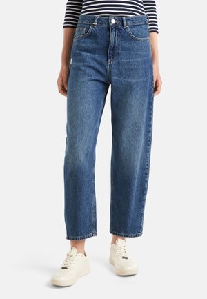 Personne portant un jean bleu taille haute à jambes droites, des baskets blanches et un haut à manches longues rayé noir et blanc, debout sur un fond blanc.