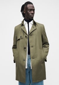 Trenci verde măsliniu cu guler decupat, două buzunare frontale și închidere cu nasturi. Purtați peste o cămașă albă și blugi albaștri. Textură netedă.