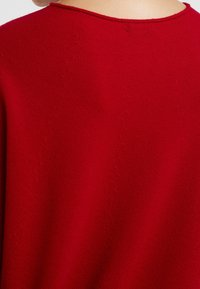 Pull rouge en tricot avec un col rond, présentant un motif texturé et une bordure côtelée au col. Tissu doux avec une finition lisse.