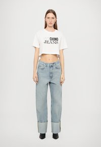 Iškirpta balta marškinė su juodu užrašu "CHINO JEANS"; šviesiai mėlynos laisvos formos džinsai su sulankstytais užtvarais ir tamsesniu akcentu ties apdaila.