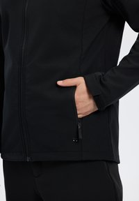 Veste softshell noire avec fermeture éclair, dotée d'une poche latérale et de poignets ajustables, fabriquée dans un matériau lisse et extensible.