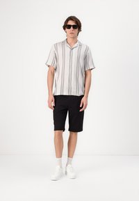 Camisa de manga corta a rayas en gris claro y blanco, pantalones cortos negros y zapatillas blancas. El modelo lleva gafas de sol negras y está de pie frente a un fondo blanco.