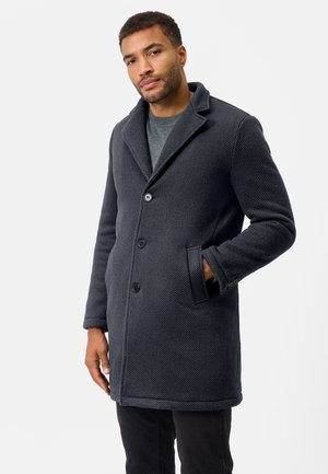 Dunkelgrauer Wollmantel mit einer strukturierten Oberfläche, Reverskragen, zwei Knöpfen und seitlichen Taschen. Das Model trägt einen grauen Strickpullover darunter.