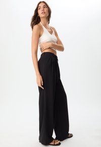 Top corto bianco con reggiseno alla halter abbinato a pantaloni neri a vita alta e gamba larga con cintura annodabile e una texture elegante. Sandali neri completano l'outfit.