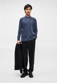 Μπλε πόλο μπλούζα με μακριά μανίκια και μαλακή υφή, με τρία κουμπιά στο λαιμό, συνδυασμένη με μαύρο παντελόνι και παπούτσια, κρατώντας μια μαύρη ζακέτα.