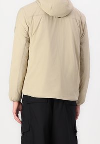 Beżowa kurtka z kapturem i elastycznymi mankietami, noszona z czarnymi spodniami typu cargo, widoczna od tyłu na osobie stojącej na jednolitym tle.