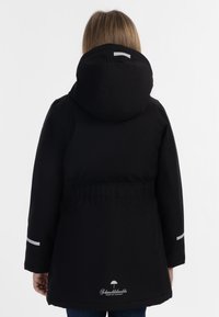 Černá parka s kapucí, s užším pasem, reflexními prvky na rukávech a logem na zadní straně, vystavená na bílém pozadí.
