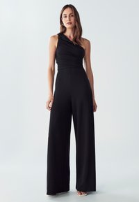 Čierny jednoramenný overal so zhrnutou látkou, širokými nohavicami, hladkou textúrou a švami. U.Style s svetlými vysokými podpätkami.