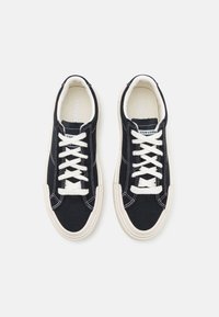 Svarta kanvasnikar med vita snören, kontrasterande vit sömnad och en krämfärgad gummisula. Rundad tå och vadderad krage.