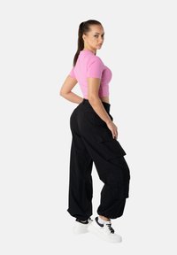 Poninhännäinen nainen, joka käyttää vaaleanpunaista lyhyttä t-paitaa, mustia cargo-housuja ja valkoisia tennareita, seisoo käsi lanteilla ja katsoo sivulle.