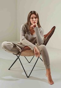 Beige geripptes Strick-Loungewear-Set mit einem V-Ausschnitt-Pullover und passender Jogginghose, sitzend in einem braunen Wildleder-Schmetterlingssessel mit schwarzem Metallgestell.