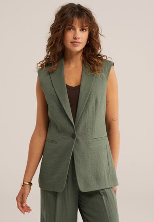 Kobieta o kręconych brązowych włosach, ubrana w bezrękawnik w kolorze oliwkowej zieleni na ciemnobrązowej bluzce oraz dopasowane oliwkowozielone spodnie, neutralne tło.