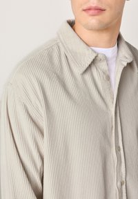 Mladić koji nosi svijetlo bež kosiranu košulju s gumbima preko bijele majice okruglog ovratnika, djelomični prikaz od ramena do usana.