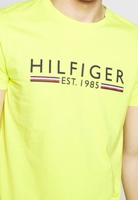 Camiseta de algodón de manga corta en amarillo neón, que presenta "HILFIGER EST. 1985" en negrita con texto negro y franjas horizontales rojas y azules.