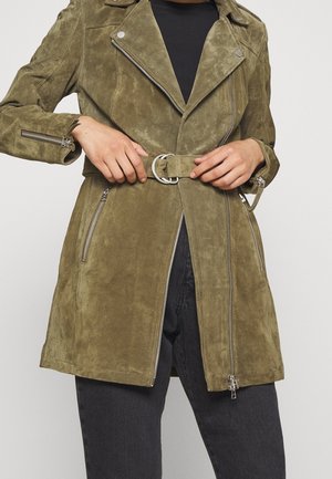 Personne attachant une ceinture sur une veste en daim vert olive avec des fermetures éclair argentées sur un top noir et un jean foncé sur un fond uni.