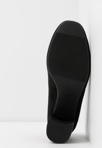 Semelle de chaussure noire avec motif de grip texturé, inclinée contre un mur et un sol blancs.
