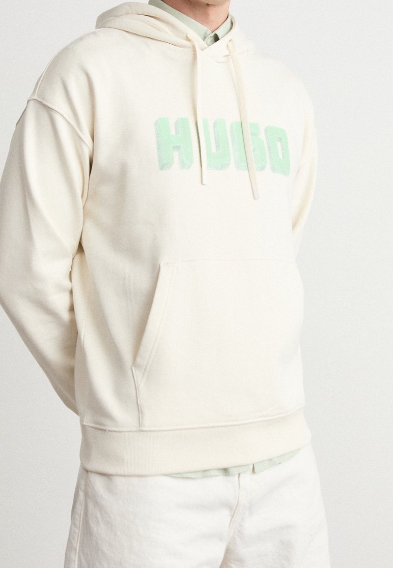 Sweat à capuche crème avec une poche avant, présentant un texte "HUGO" en relief vert menthe. Tissu léger, coupe ample, capuche avec cordon de serrage et poignets côtelés.