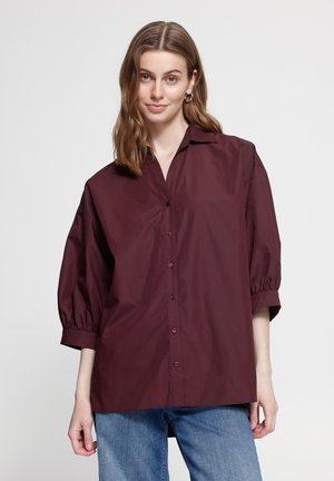 Giovane donna con capelli castani ondulati indossa una camicia scura bordeaux con bottoni e maniche a sbuffo tre quarti e jeans blu, in piedi davanti a uno sfondo semplice.