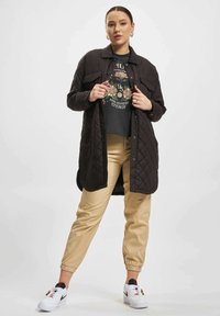 Quilted svart jakke over en blomstret grafisk skjorte, beige bukser med elastiske mansjetter, og hvite sneakers med svarte detaljer. Modellen poserer selvsikkert.