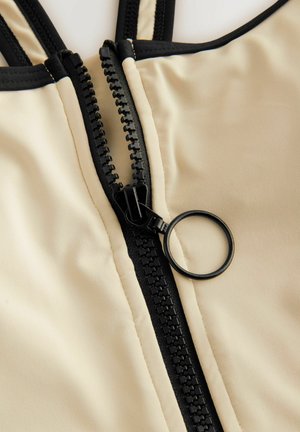 Tissu beige avec une fermeture éclair noire verticale, dotée d'un tirette circulaire et d'une couture d'accent noir près des bords.