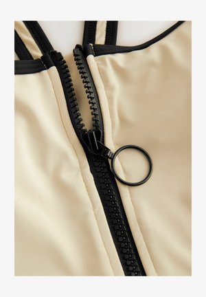 Tissu beige avec une fermeture éclair noire verticale, dotée d'un tirette circulaire et d'une couture d'accent noir près des bords.