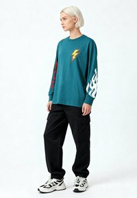 Personne aux cheveux blonds courts portant un t-shirt à manches longues bleu sarcelle avec des graphismes d'éclair et de flamme, un pantalon cargo noir et des baskets blanches.