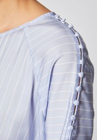 Blusa azzurro chiaro con sottili righe bianche. Presenta un colletto rotondo e dettagli di bottone lungo la cucitura della spalla. Tessuto liscio e morbido.