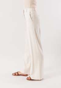 Pantaloni a gamba larga in tessuto beige chiaro, con pince, vita sartoriale e una texture liscia. Abbinati a sandali scivolati marroni.