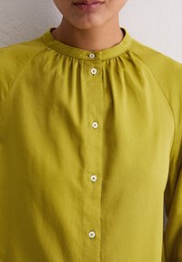 Persoon die een knopen olijfgroene blouse met een ronde kraag draagt tegen een effen lichte muur. 